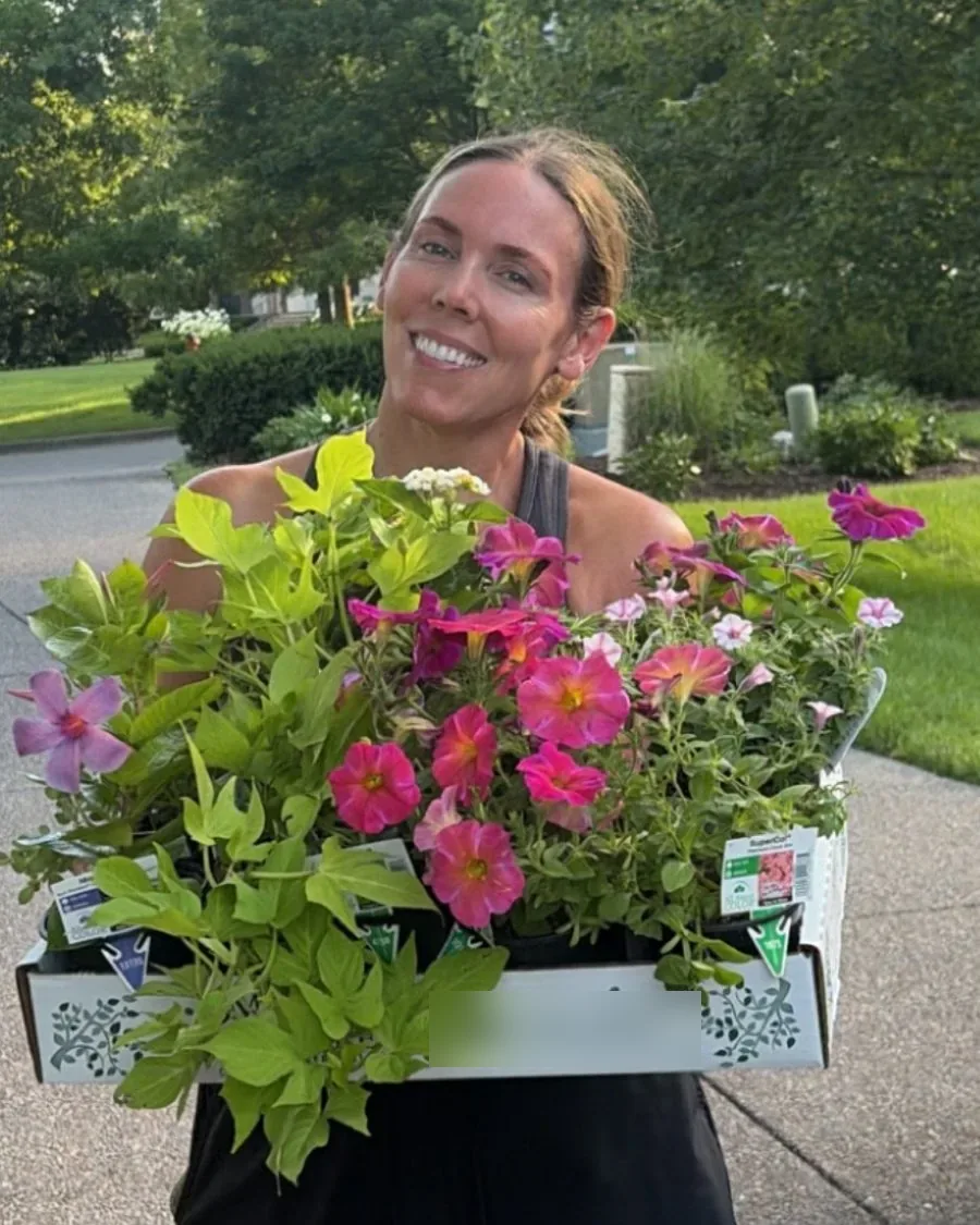 Carrie Ball, owner of Porch + Bloom Co., smiling and carrying a tray of pink petunias and greenery