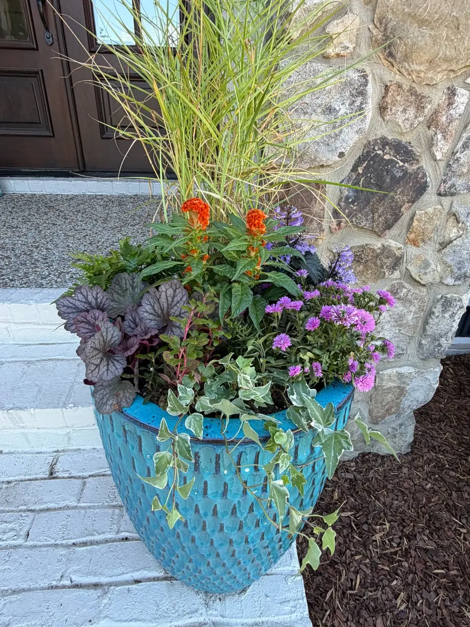 Blue glazed planter with ornamental grass, orange celosia, purple asters, and heuchera beside a stone wall.